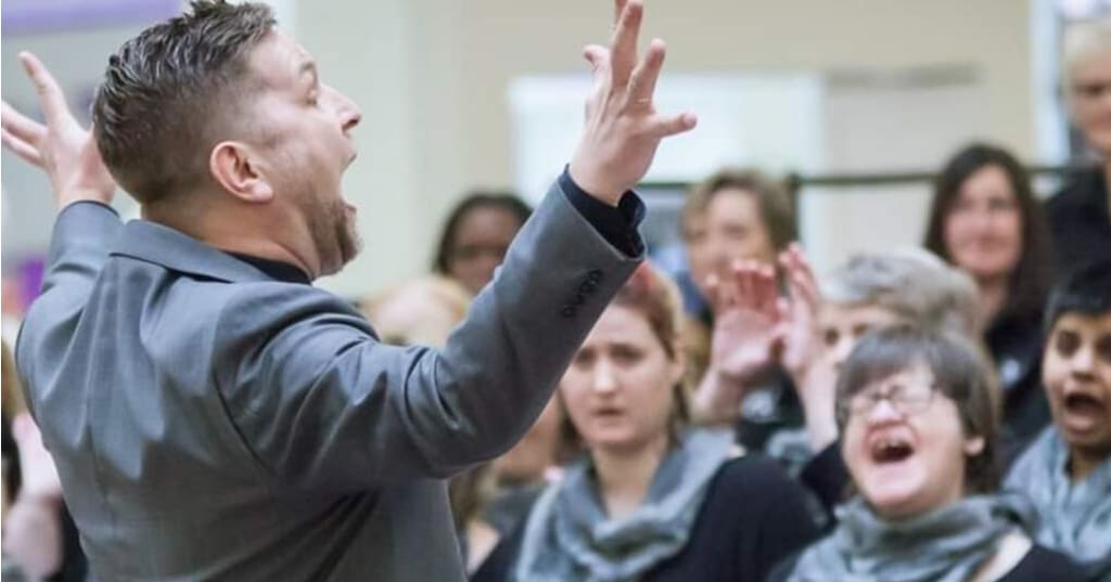 Man in focus singing and directing a choir in the background slightly out of focus.