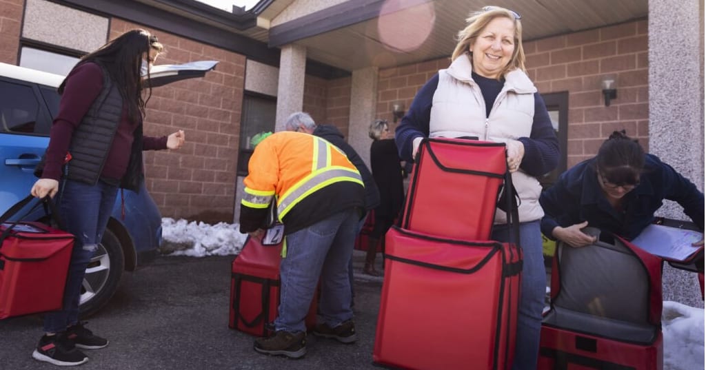 Smiling volunteers carrying out meals in warm boxes for delivery.