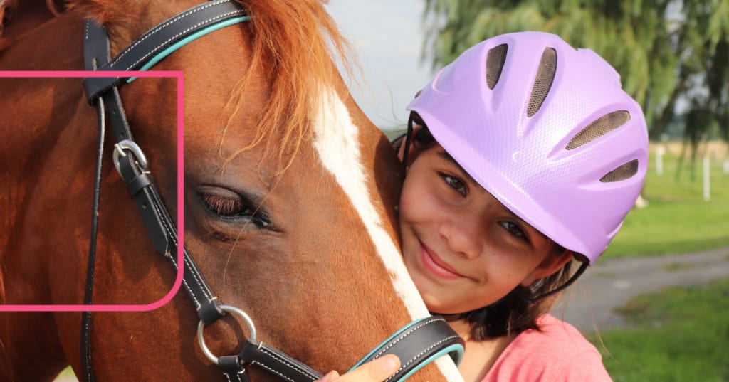Young girl wearing a pink helmet and hugging a horse's face
