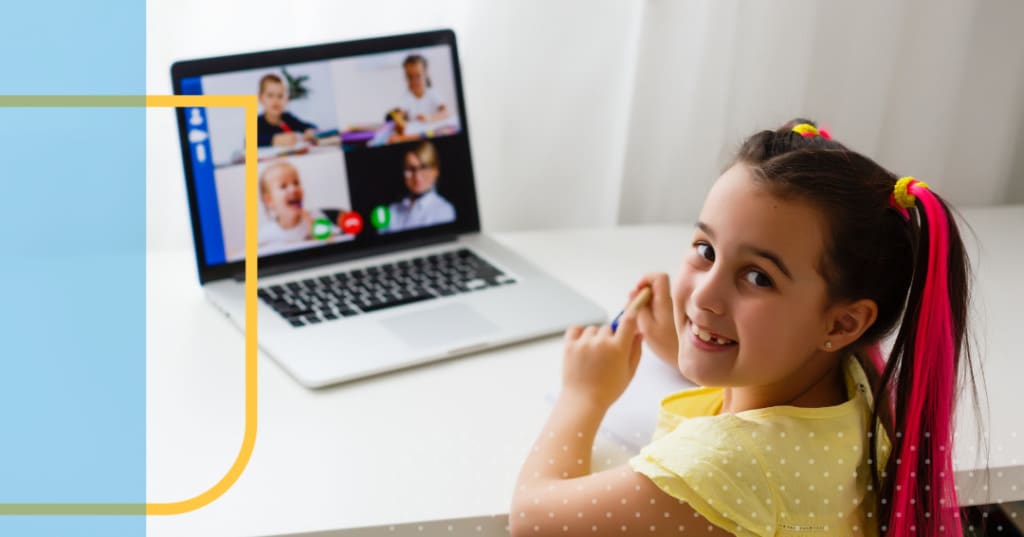 Little girl smiling with a laptop in on the table behind her showing a video chat.