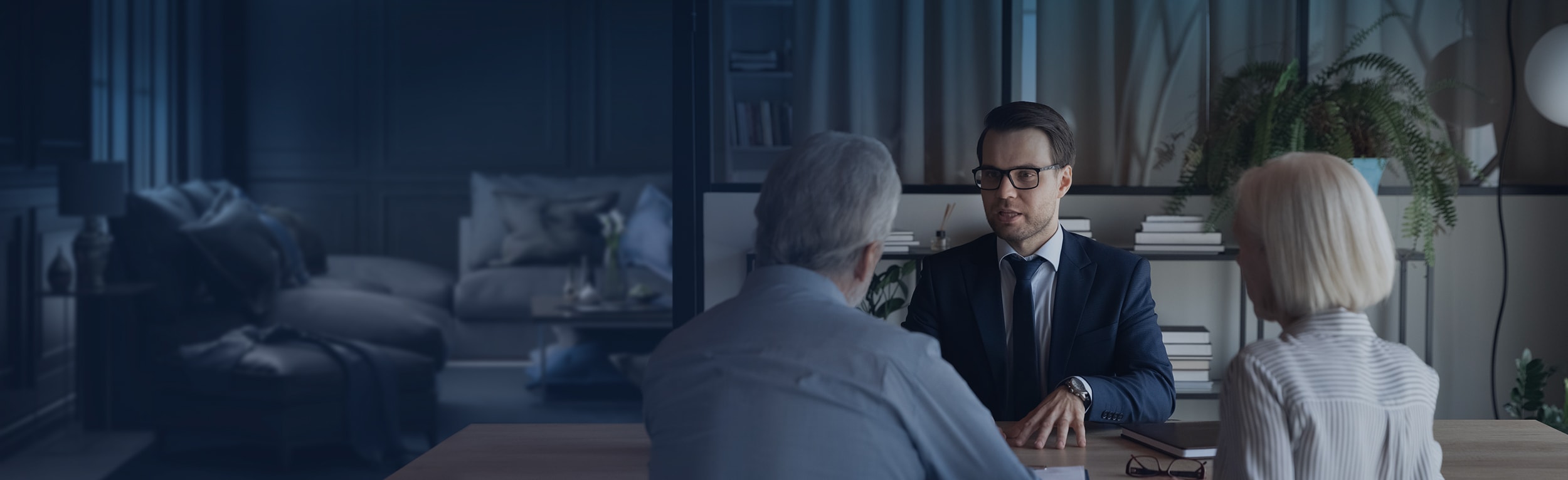 A man in a suit is sitting behind a desk talking to an older couple.