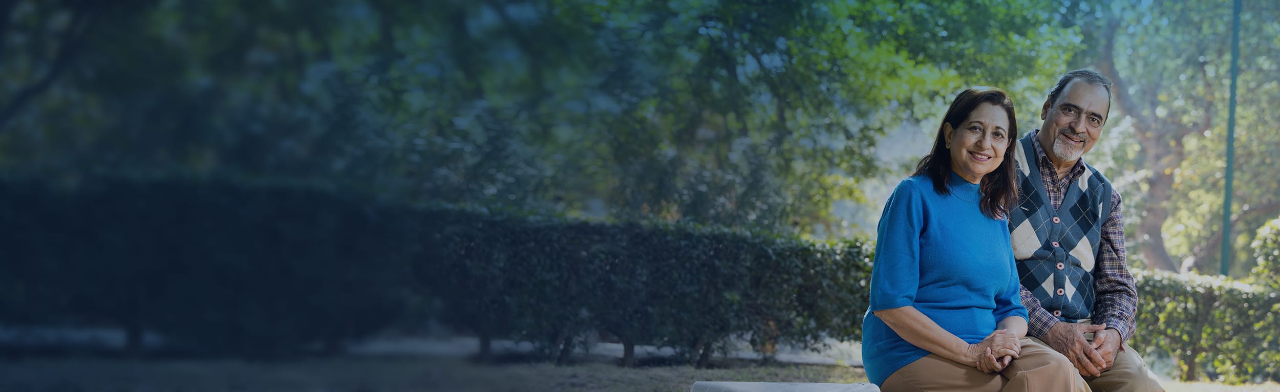 A couple os sitting on a bench outside by a hedge.